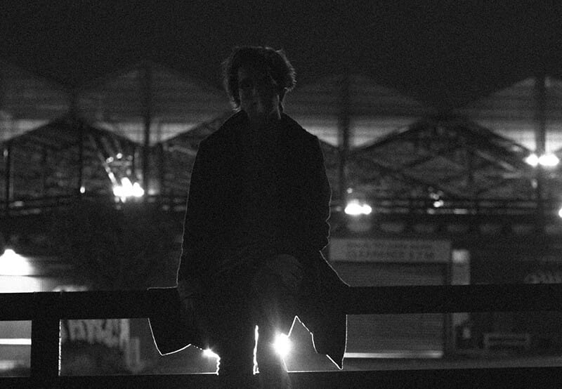 A black and white photo of a person standing on a railing at night, looking as if they are about to Camikaze off.