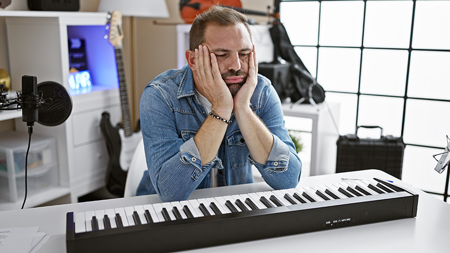 A frustrated musician sitting at a keyboard in a modern home studio, resting his face in his hands, indicating writer's block or creative frustration. 